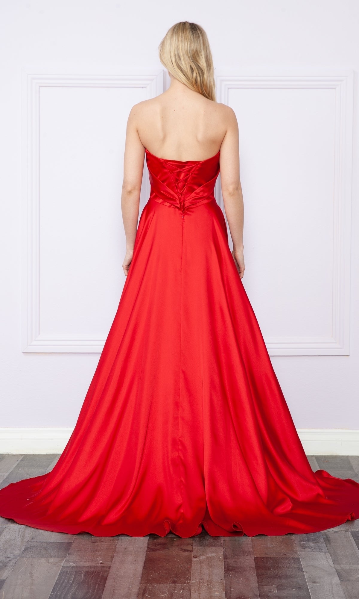 A woman faces away, wearing a strapless long A-line prom dress with pockets, fitted bodice, lace-up back, and flowing skirt with a subtle side slit, standing against a pale wall with decorative molding.